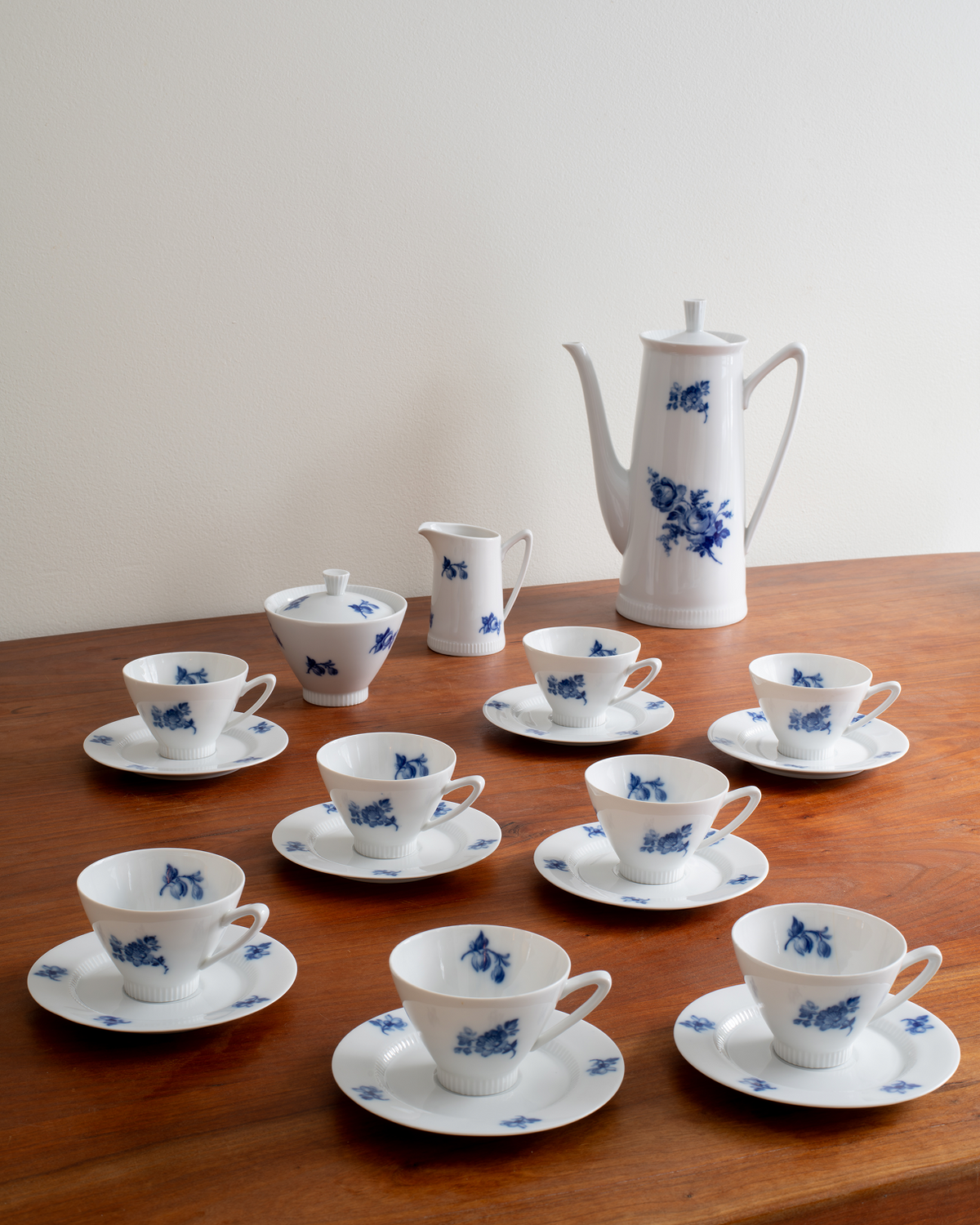 A white porcelain tea or coffee set with blue floral motifs, including a teapot, creamer, sugar bowl, and multiple cups with saucers, arranged on a wooden table against a plain light wall.