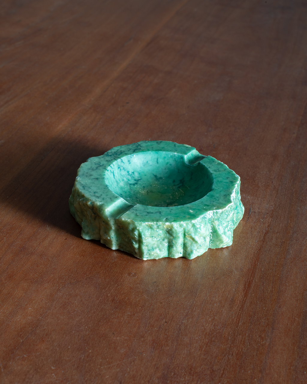 A small round green stone ashtray with a textured surface and three recessed ash tips, placed on a dark brown wooden tabletop.