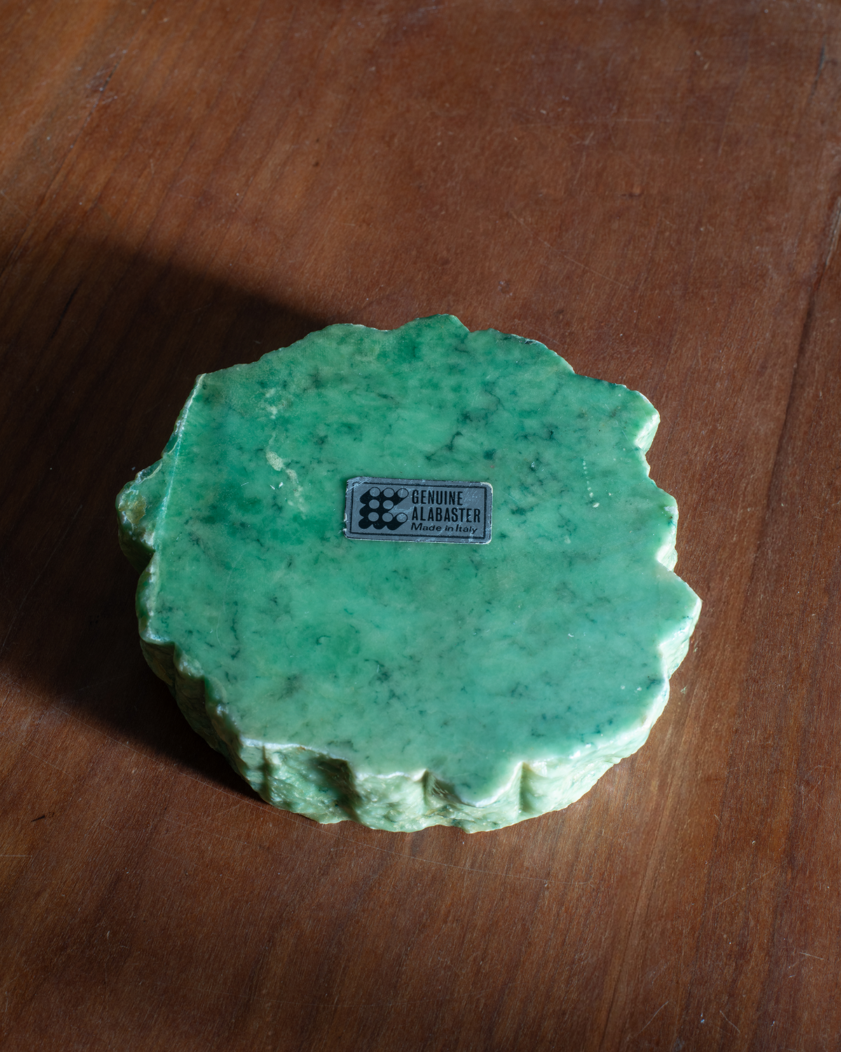 A round, scalloped-edge green alabaster ashtray with a small central label, placed on a brown wooden surface.