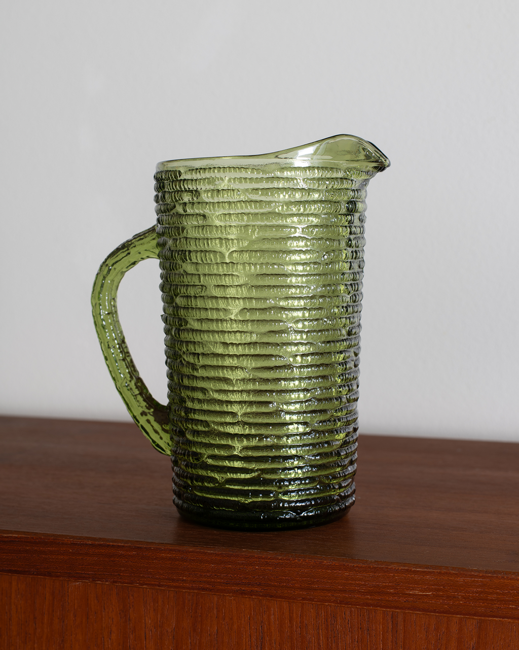 An olive green ribbed glass pitcher with a handle and pouring spout, shown sitting on a wooden surface against a plain light gray wall.