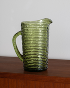 An olive green ribbed glass pitcher with a handle and pouring spout, shown sitting on a wooden surface against a plain light gray wall.