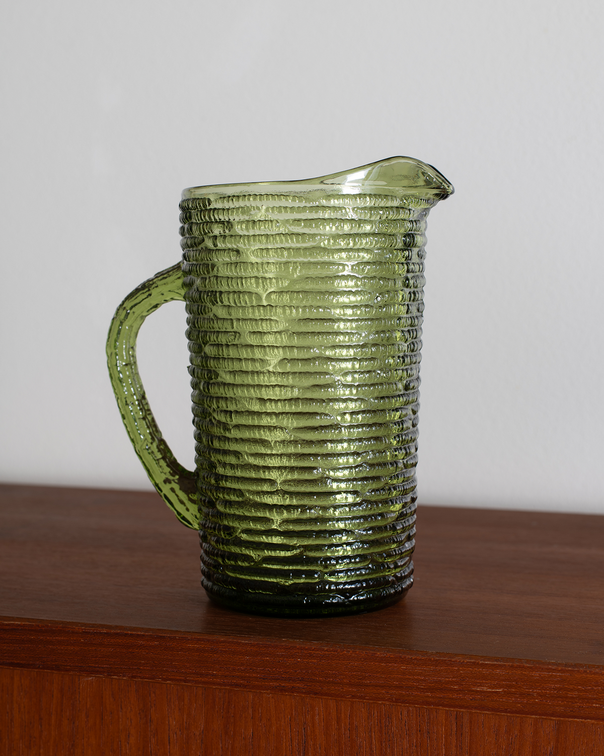 An olive green ribbed glass pitcher with a handle and pouring spout, shown sitting on a wooden surface against a plain light gray wall.