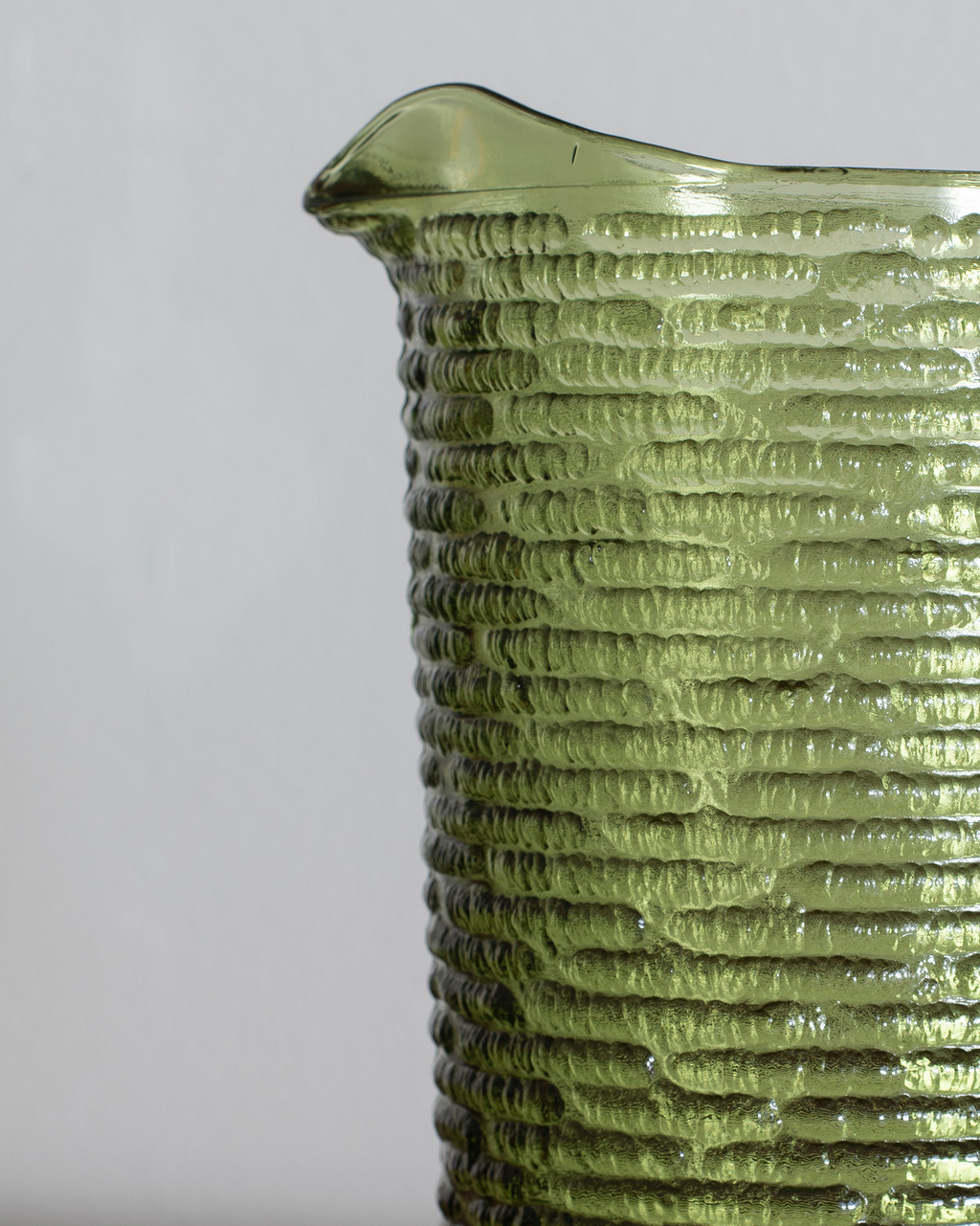 Close-up of a textured olive green glass pitcher with a ribbed pattern and a small pouring spout, shown against a neutral light gray background.