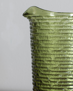 Close-up of a textured olive green glass pitcher with a ribbed pattern and a small pouring spout, shown against a neutral light gray background.