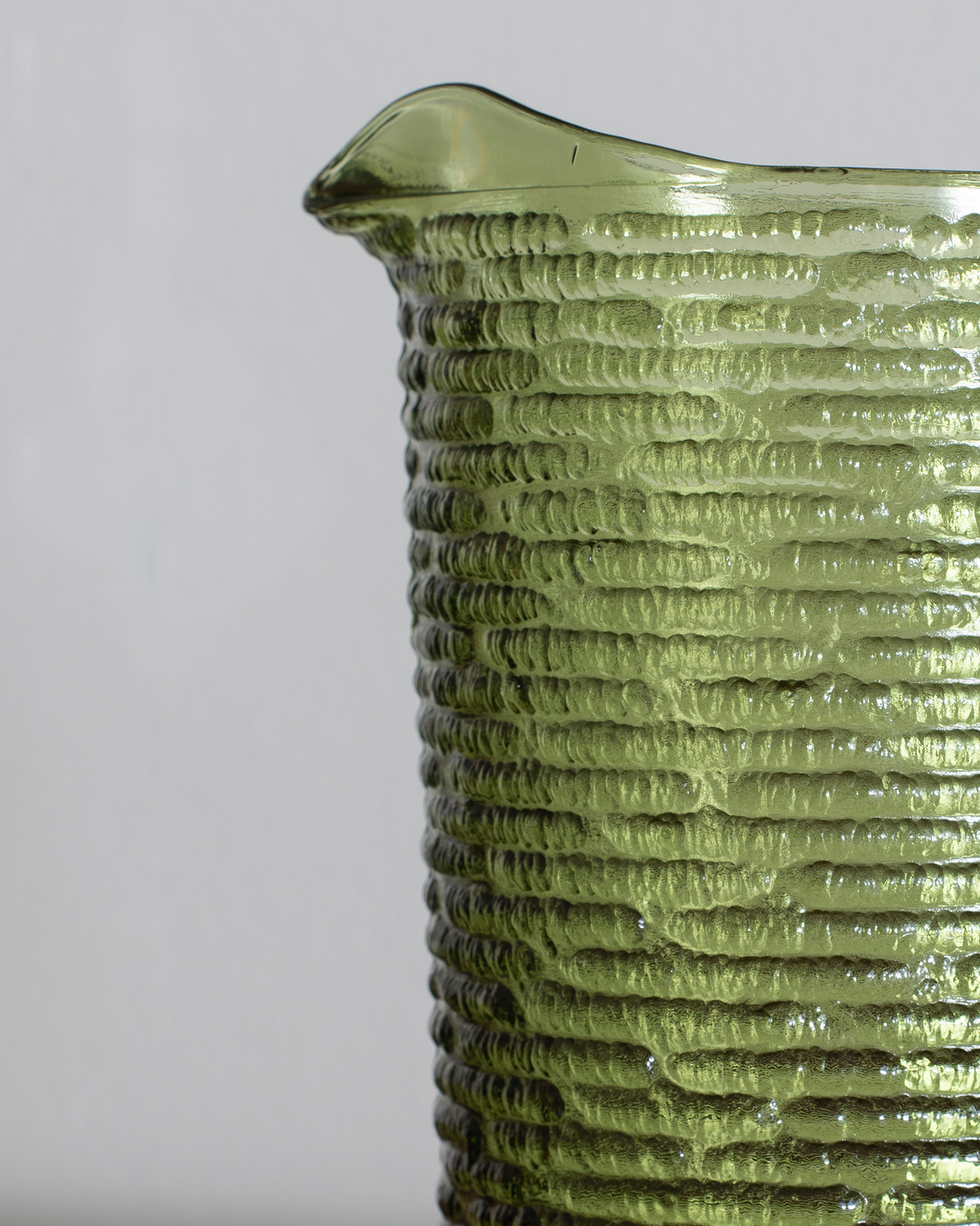 Close-up of a textured olive green glass pitcher with a ribbed pattern and a small pouring spout, shown against a neutral light gray background.