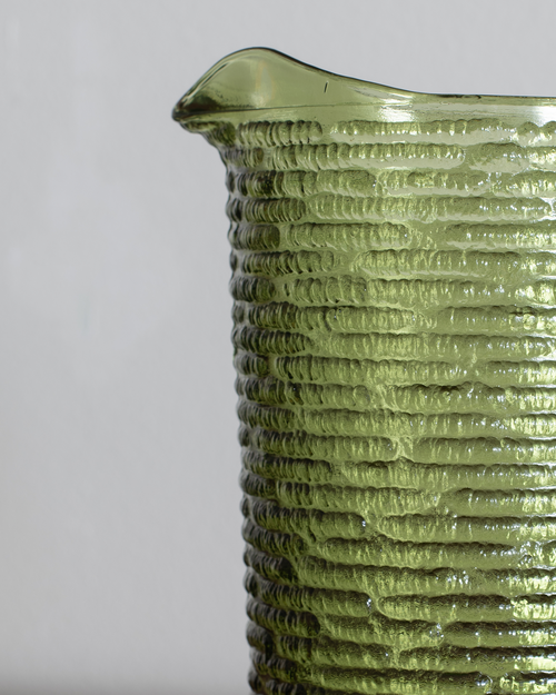 Close-up of a textured olive green glass pitcher with a ribbed pattern and a small pouring spout, shown against a neutral light gray background.