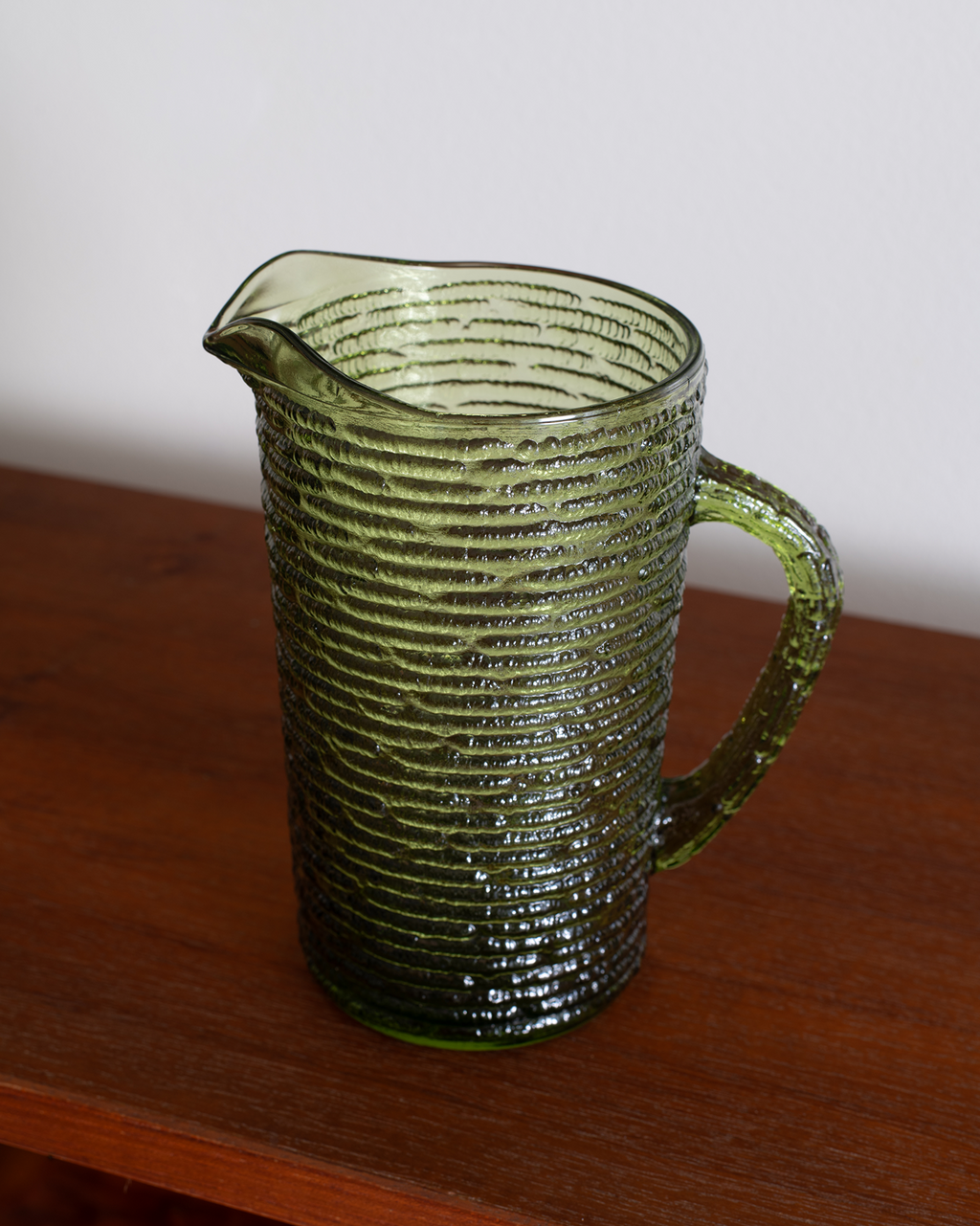 A vintage green ribbed glass pitcher with a handle and pouring spout, resting on a wooden table against a plain light gray wall.