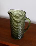 A vintage green ribbed glass pitcher with a handle and pouring spout, resting on a wooden table against a plain light gray wall.