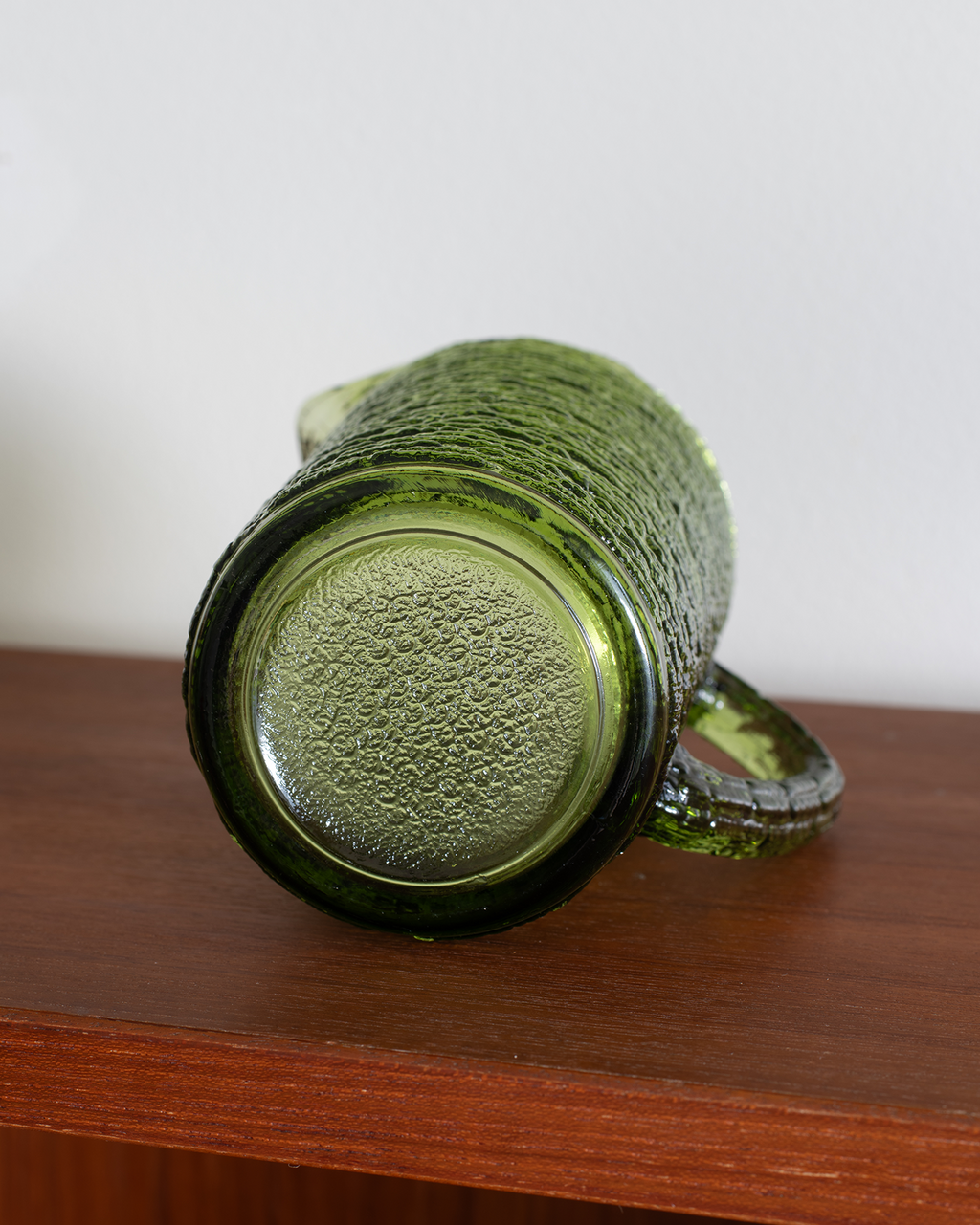 A vintage green textured glass mug with a ribbed handle, shown lying on its side on a wooden shelf against a plain white wall.