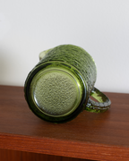 A vintage green textured glass mug with a ribbed handle, shown lying on its side on a wooden shelf against a plain white wall.
