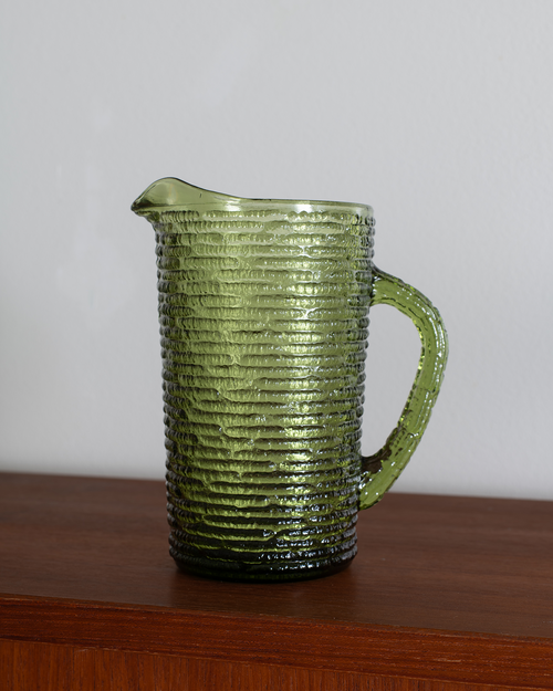 A textured olive green glass pitcher with a ribbed pattern and a curved handle, sitting on a wooden surface against a plain light gray wall.