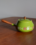 A bright green enamel teapot with a wooden handle and lid knob, decorated with white and pink floral patterns, resting on a wooden table against a gray background.