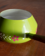 A lime green enamel saucepan with a dark green wooden handle, decorated with pink and white floral patterns, resting on a wooden table.