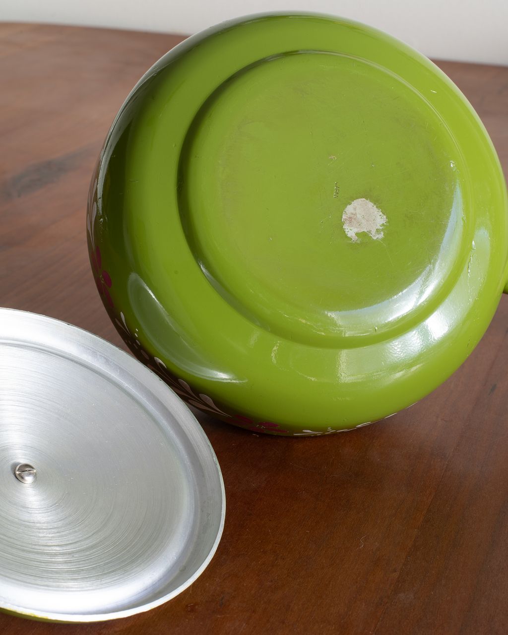 A close-up of a glossy lime green ceramic pot or pan base with a detached silver metal lid resting on a wooden tabletop.