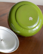 A close-up of a glossy lime green ceramic pot or pan base with a detached silver metal lid resting on a wooden tabletop.