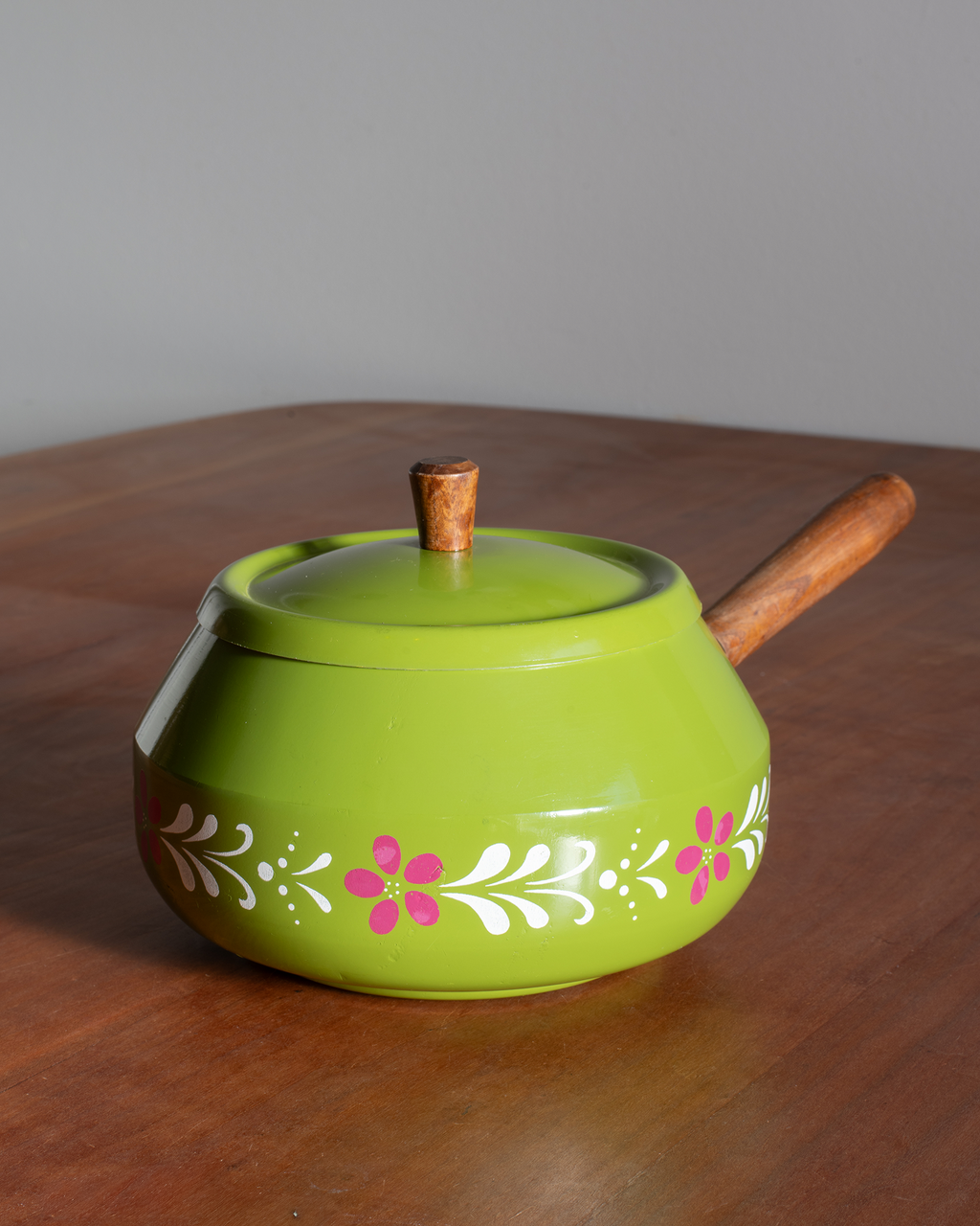 A bright green ceramic teapot with a wooden handle and lid knob, decorated with white and pink floral patterns around the lower half, sitting on a wooden table against a gray background.