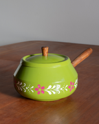 A bright green ceramic teapot with a wooden handle and lid knob, decorated with white and pink floral patterns around the lower half, sitting on a wooden table against a gray background.