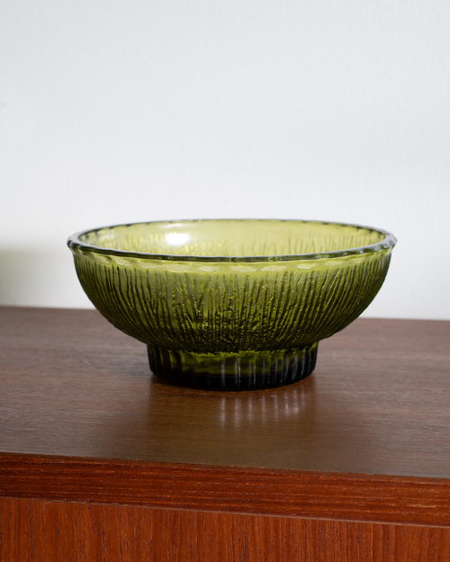 An olive green ribbed glass bowl with a flared rim and pedestal base, resting on a polished wooden surface against a plain white wall.