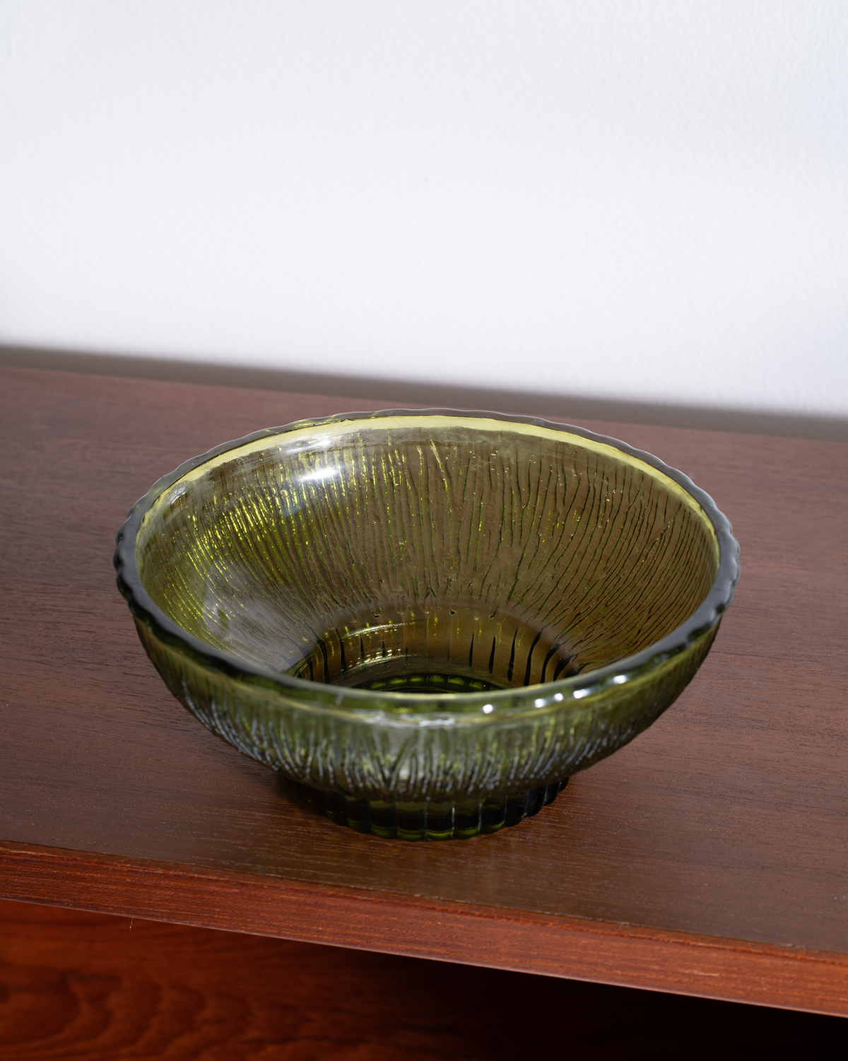 A textured olive green glass bowl with a ribbed base, resting on a dark wooden shelf against a plain white wall.