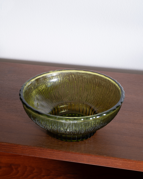 A textured olive green glass bowl with a ribbed base, resting on a dark wooden shelf against a plain white wall.