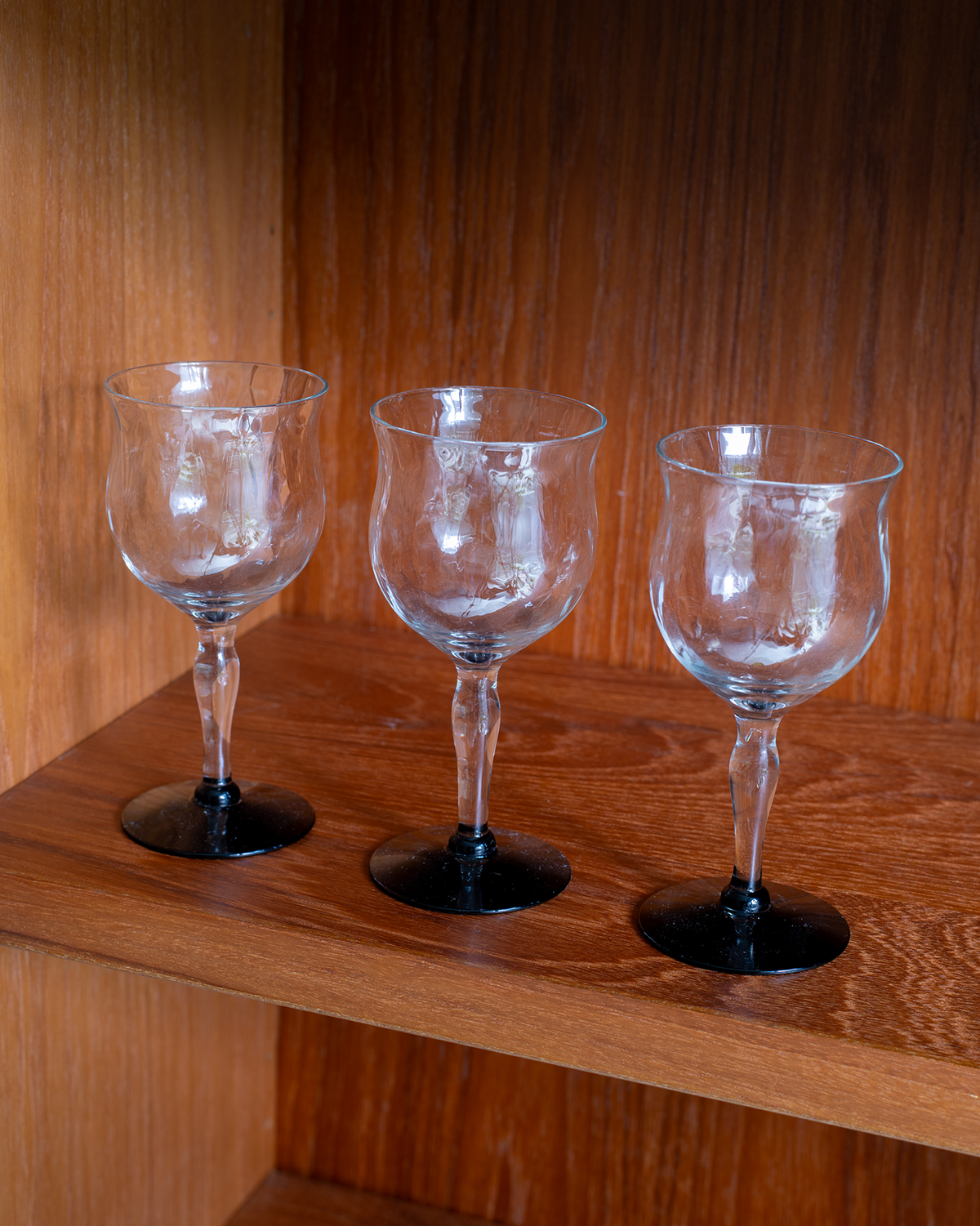 Three clear glass wine glasses with black circular bases, arranged side by side on a wooden shelf against a wooden background.