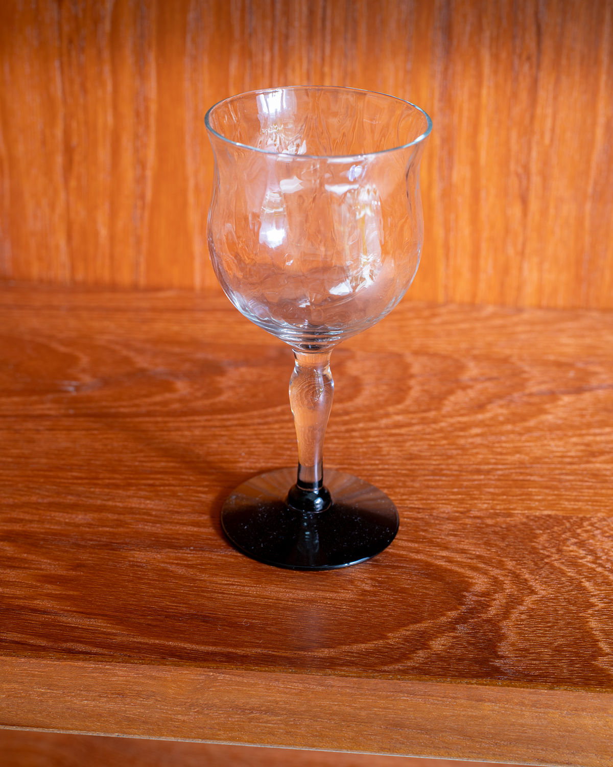 A clear glass goblet with a black circular base, standing on a polished wooden surface against a wooden background.