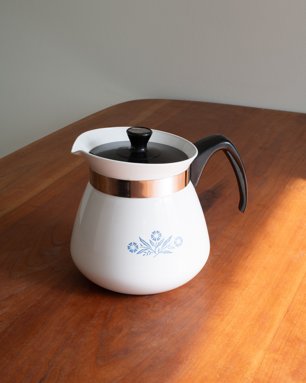 A white enamel coffee pot with a black handle and lid, featuring a copper-colored metallic band and a blue floral pattern on the front, sitting on a wooden table.