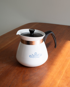 A white enamel coffee pot with a black handle and lid, featuring a copper-colored metallic band and a blue floral pattern on the front, sitting on a wooden table.