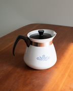 A vintage-style white coffee pot with a black handle and lid, featuring a metallic copper band and a blue floral pattern on the front, sitting on a wooden table.
