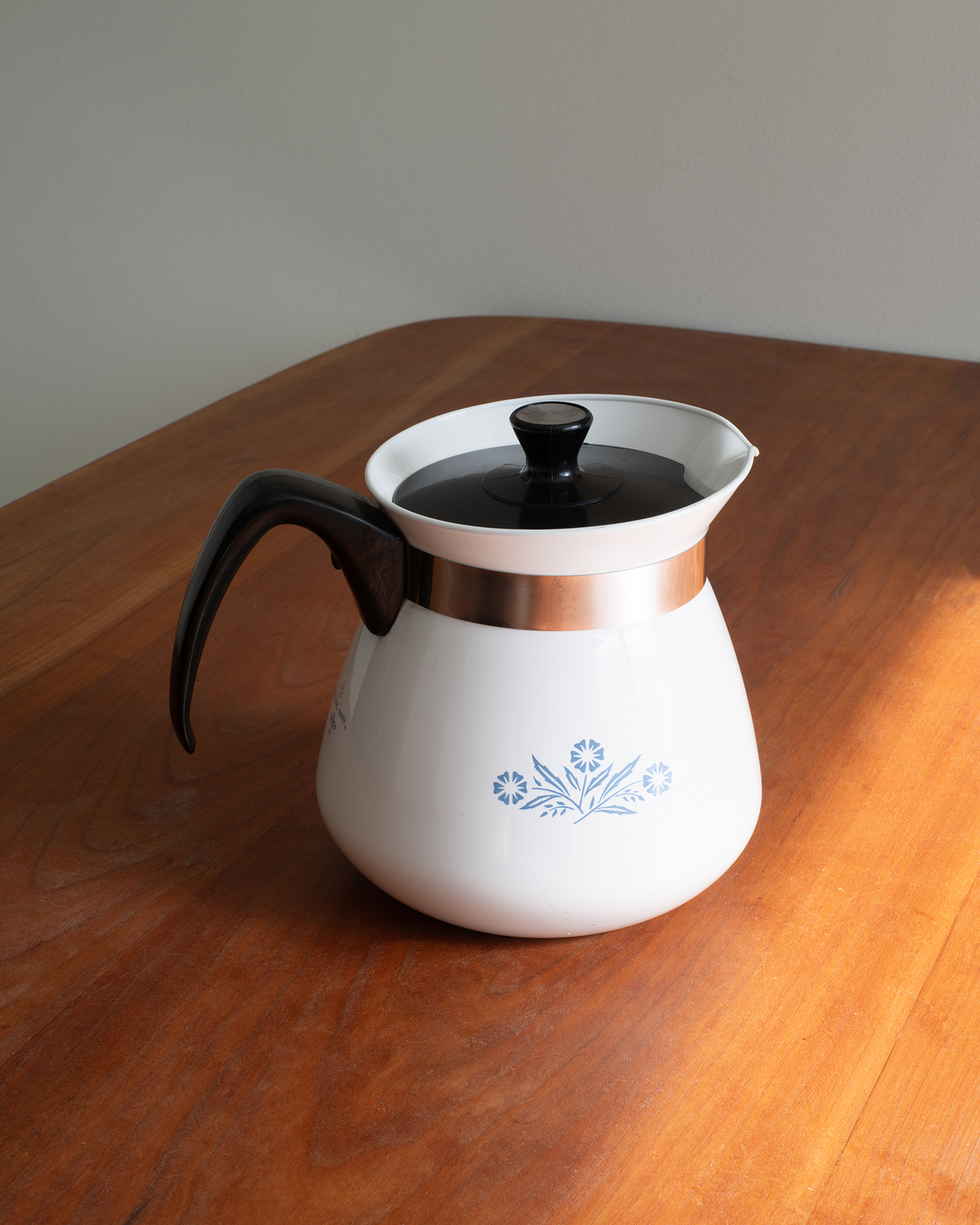 A vintage-style white coffee pot with a black handle and lid, featuring a metallic copper band and a blue floral pattern on the front, sitting on a wooden table.