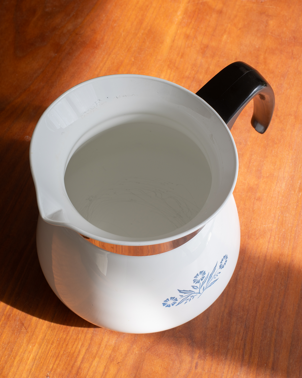 A white ceramic pitcher with a black handle and blue floral decoration, filled with water and photographed on a warm wooden tabletop.