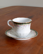 A vintage white porcelain teacup and saucer set with delicate floral patterns, a gold rim, and a green band, displayed on a wooden table.