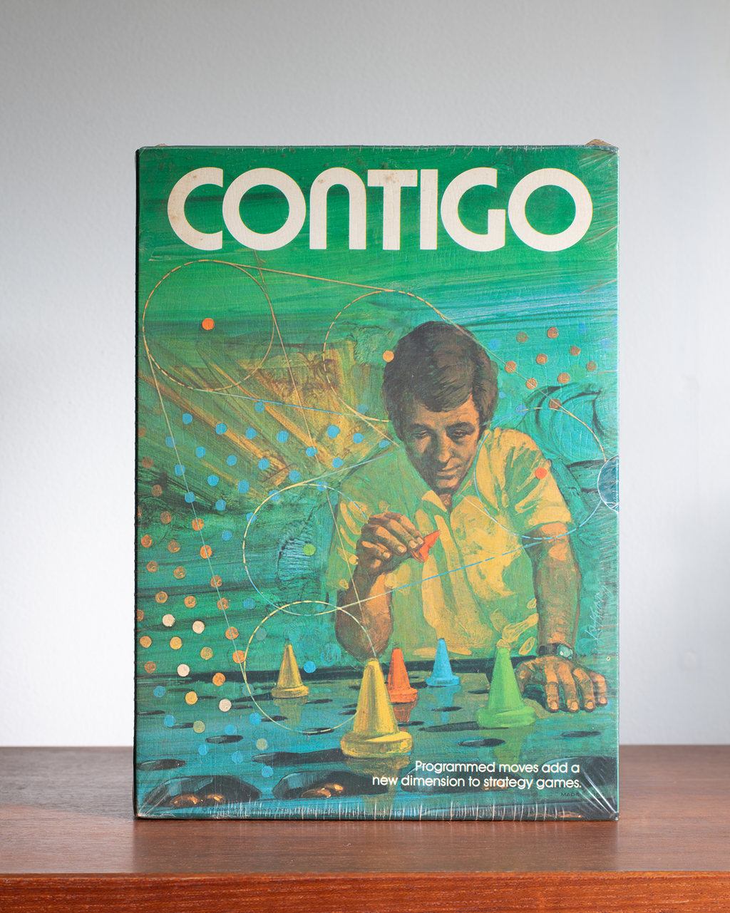 Vintage Contigo board game box on a wooden surface against a light gray background. The box features a retro illustration of a man playing with colorful geometric pieces on a teal background, with the brand name 'CONTIGO' in large white letters at the top.