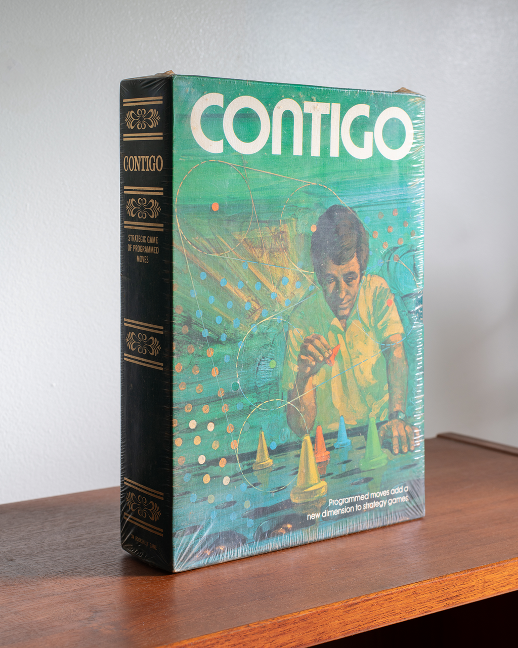 A vintage hardcover book titled 'CONTIGO' standing upright on a wooden shelf against a plain light wall. The front cover is teal-green with a retro illustration of a child and colorful geometric shapes, while the spine is black with gold lettering.