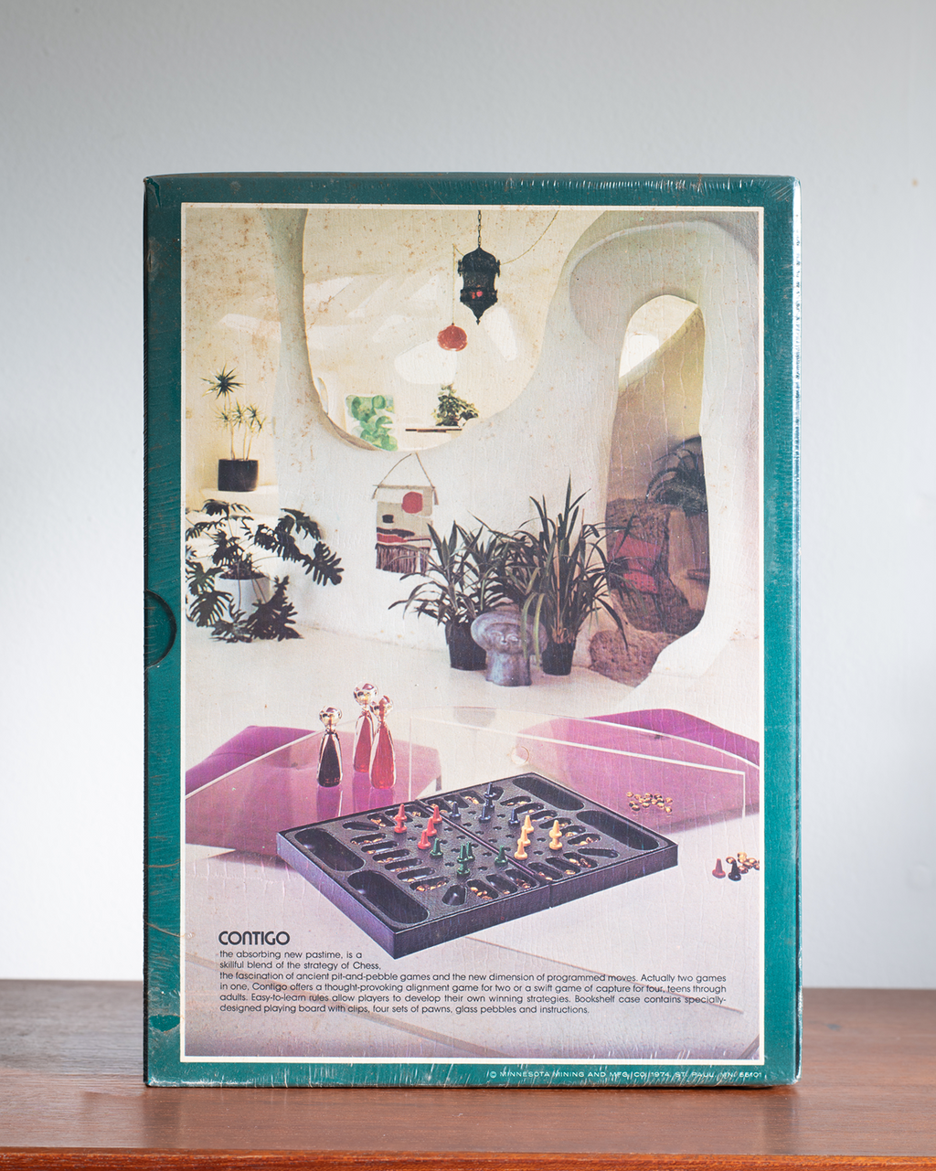 A vintage board game box for 'Contigo' is shown standing upright on a wooden surface against a light gray wall. The box features a teal border and a cover illustration depicting a retro-futuristic interior with pink lounge chairs, potted plants, and the game board in the foreground.