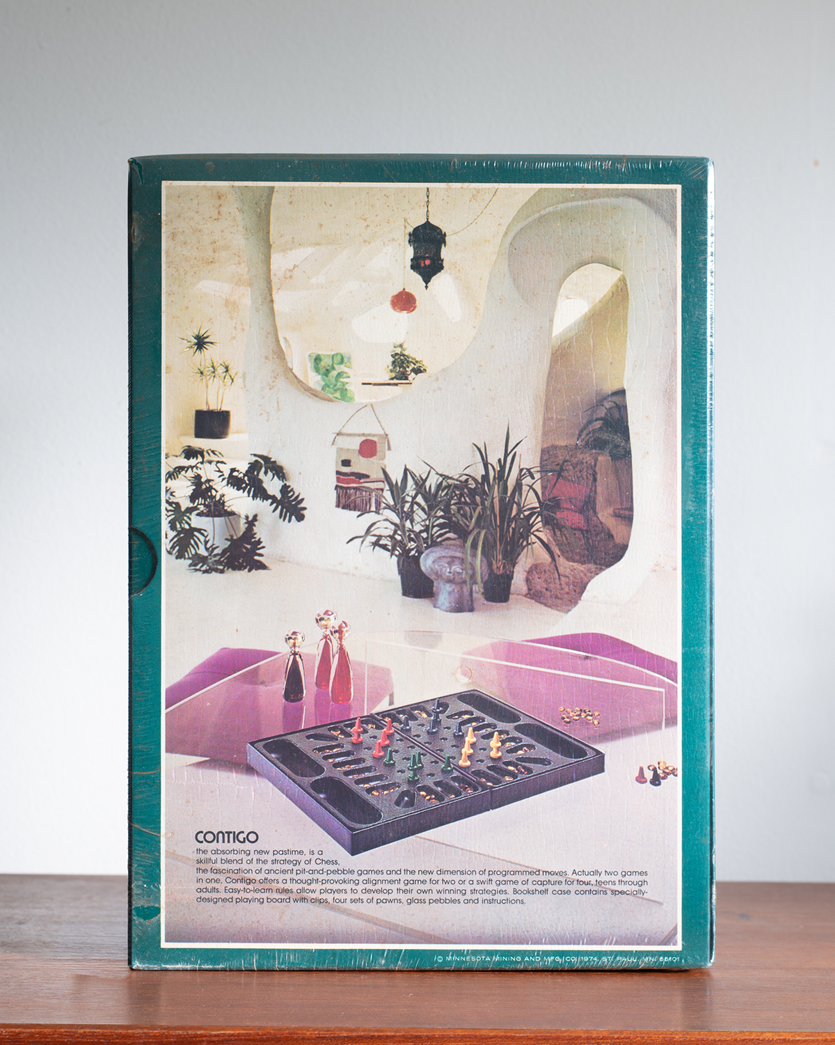 A vintage board game box for 'Contigo' is shown standing upright on a wooden surface against a light gray wall. The box features a teal border and a cover illustration depicting a retro-futuristic interior with pink lounge chairs, potted plants, and the game board in the foreground.