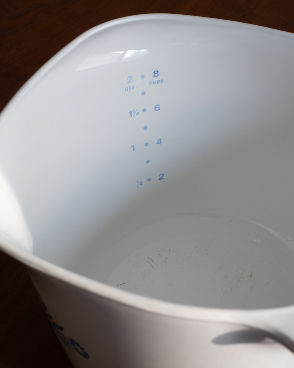 Close-up of the inside of a white measuring cup with blue printed measurement markings, photographed on a dark wooden surface.