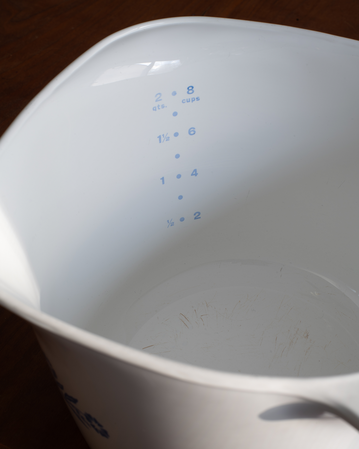 Close-up of the inside of a white measuring cup with blue printed measurement markings, photographed on a dark wooden surface.