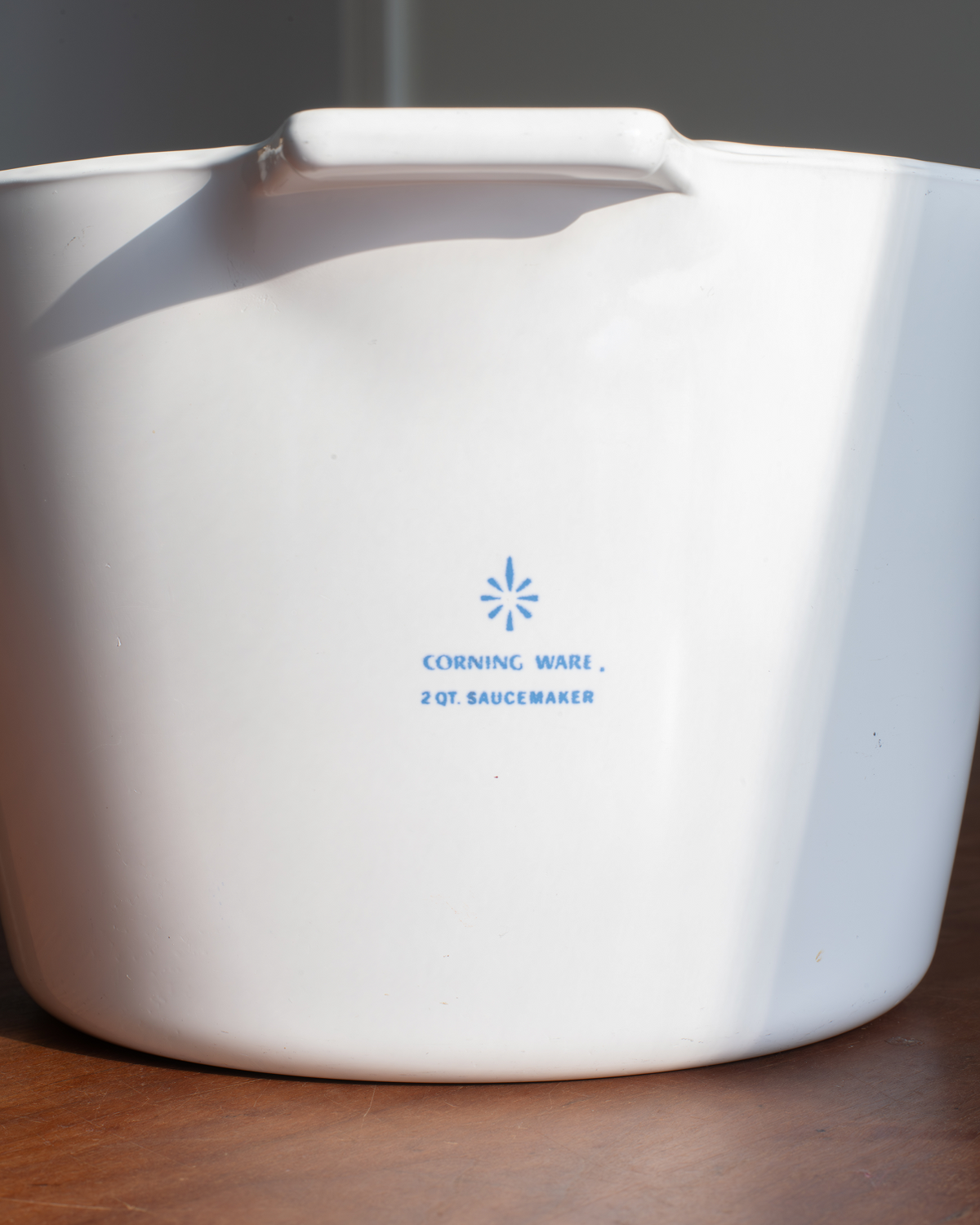 A white Corning 2 Qt Saucemaker ceramic pot with blue printed text and logo, sitting on a wooden surface with a dark background.