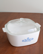 A white glass casserole dish with a matching glass lid and a blue floral pattern, sitting on a wooden table against a light wall.