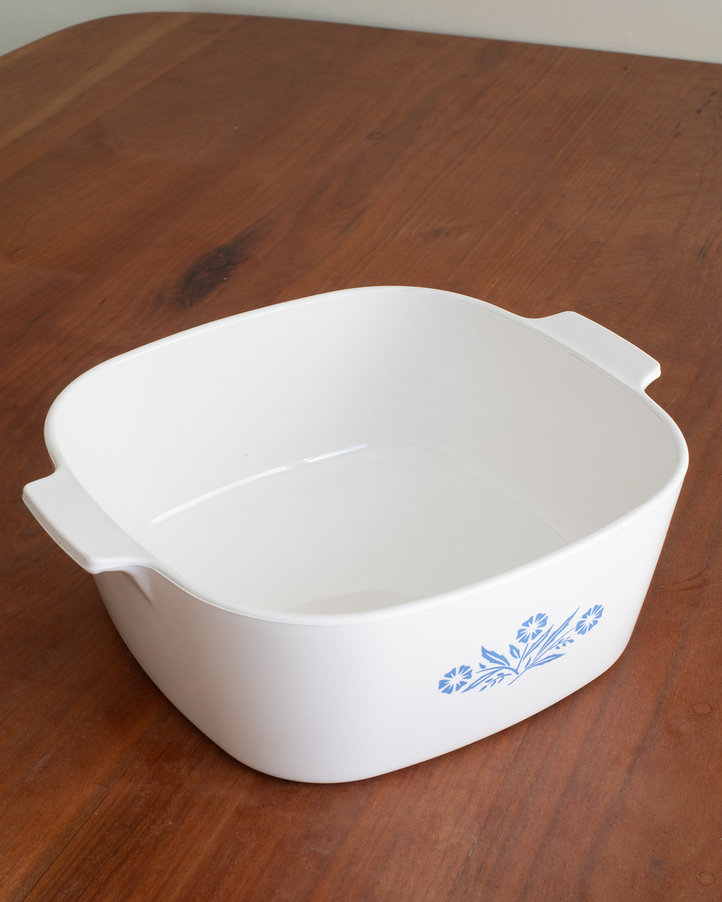 A white square ceramic casserole dish with side handles and a blue floral pattern on the front, shown on a wooden tabletop.