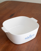 A white square ceramic casserole dish with side handles and a blue floral pattern on the front, shown on a wooden tabletop.