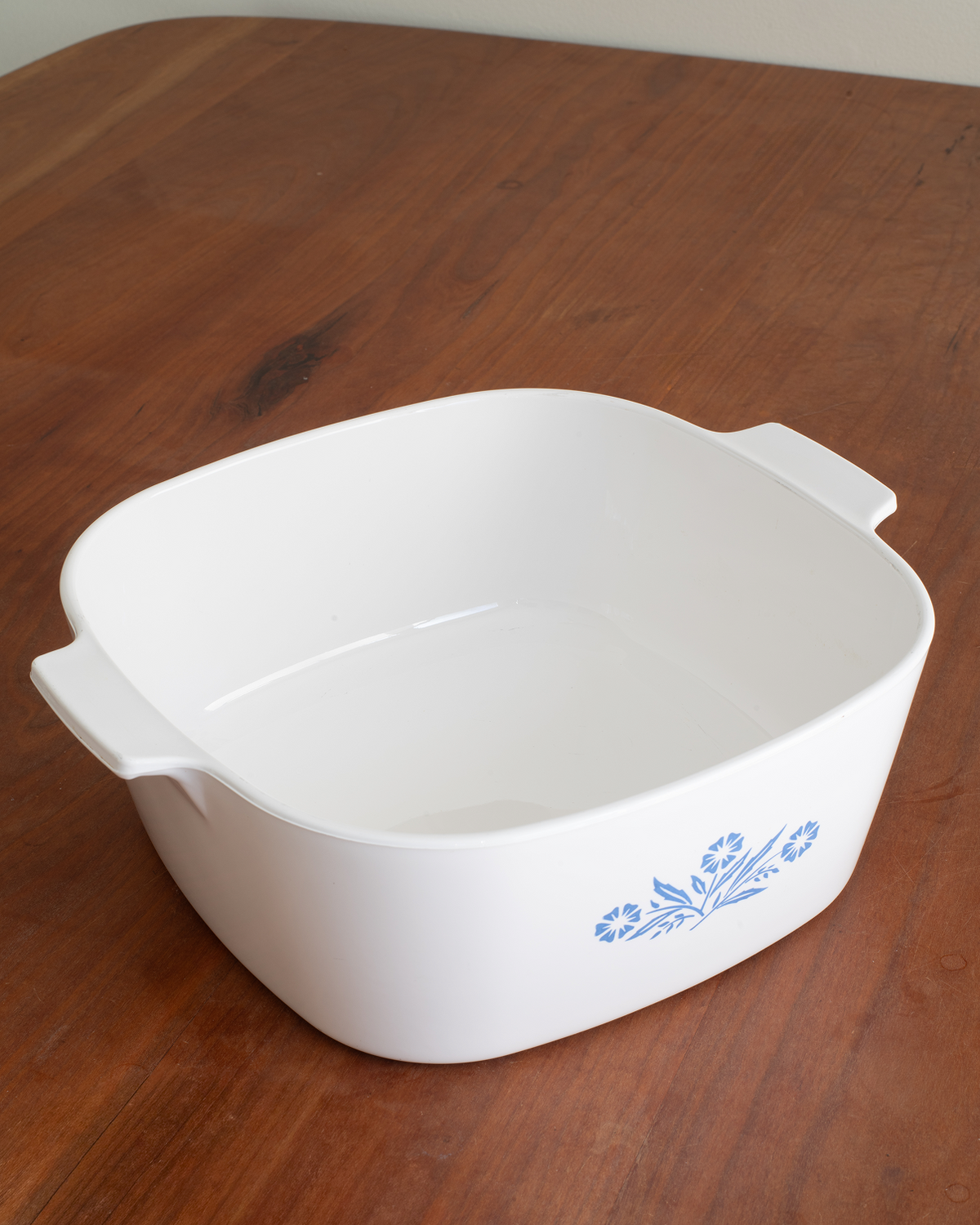 A white square ceramic casserole dish with side handles and a blue floral pattern on the front, shown on a wooden tabletop.