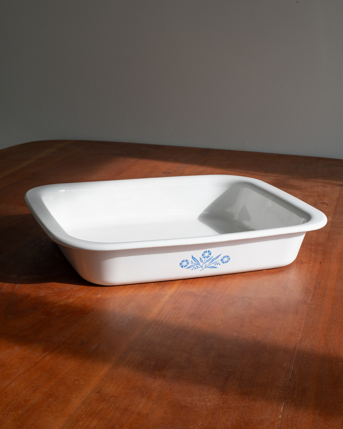 A white rectangular enamel baking dish with rounded corners and a blue floral pattern near the front, resting on a warm brown wooden table against a neutral gray background.