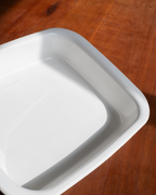 A white square ceramic baking dish with rounded corners, shown from an elevated angle on a warm brown wooden surface.