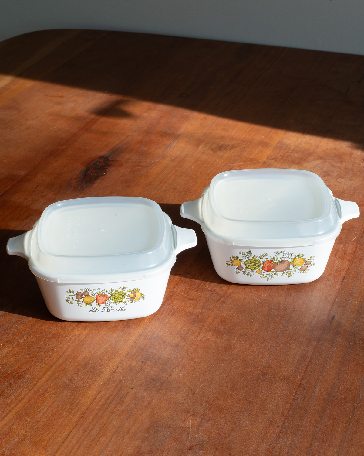 Two white casserole dishes with lids, featuring the classic Multicolor Fruit border pattern, displayed on a wooden table in natural sunlight.