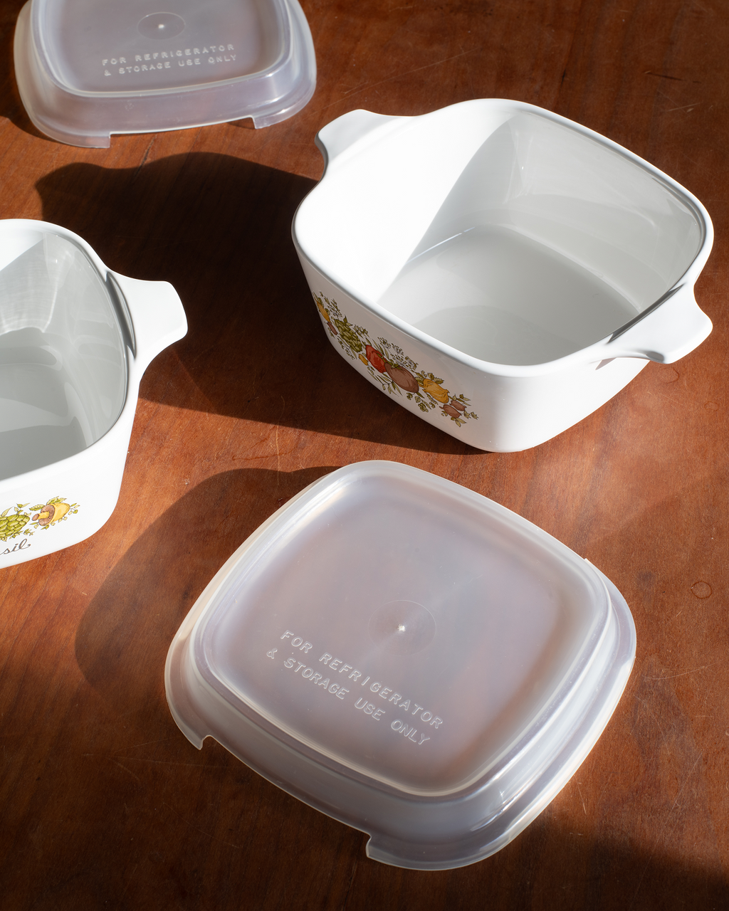 White ceramic double-compartment food storage containers with handles and translucent plastic lids, arranged on a warm brown wooden surface.
