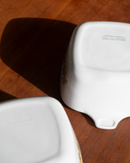 Two white ceramic baking dishes shown upside down on a wooden surface, displaying their non-stick coated interiors and printed safety markings.