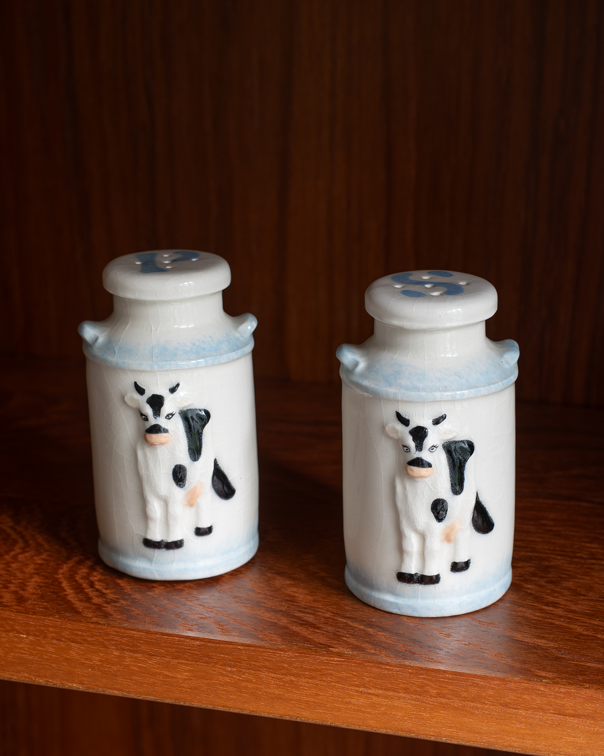 Two small ceramic salt and pepper shakers with cow designs, sitting on a wooden shelf against a dark wood background.