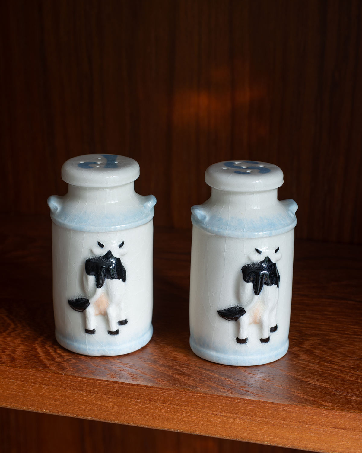 Two white ceramic salt and pepper shakers with blue rims and black-and-white cow figures, displayed on a wooden shelf against a dark wood background.